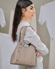 A woman in a white shirt carries a taupe leather shoulder bag with a structured slouchy shape and a small silver logo on the bottom right, set against a background of hanging white shirts.