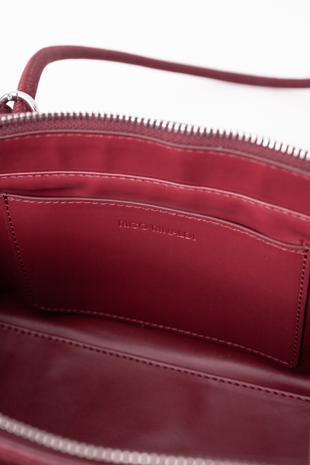 Close-up of the interior of a burgundy leather bag, showing a zippered opening, interior pocket, and debossed 'RICO RINALDI' branding on the lining.