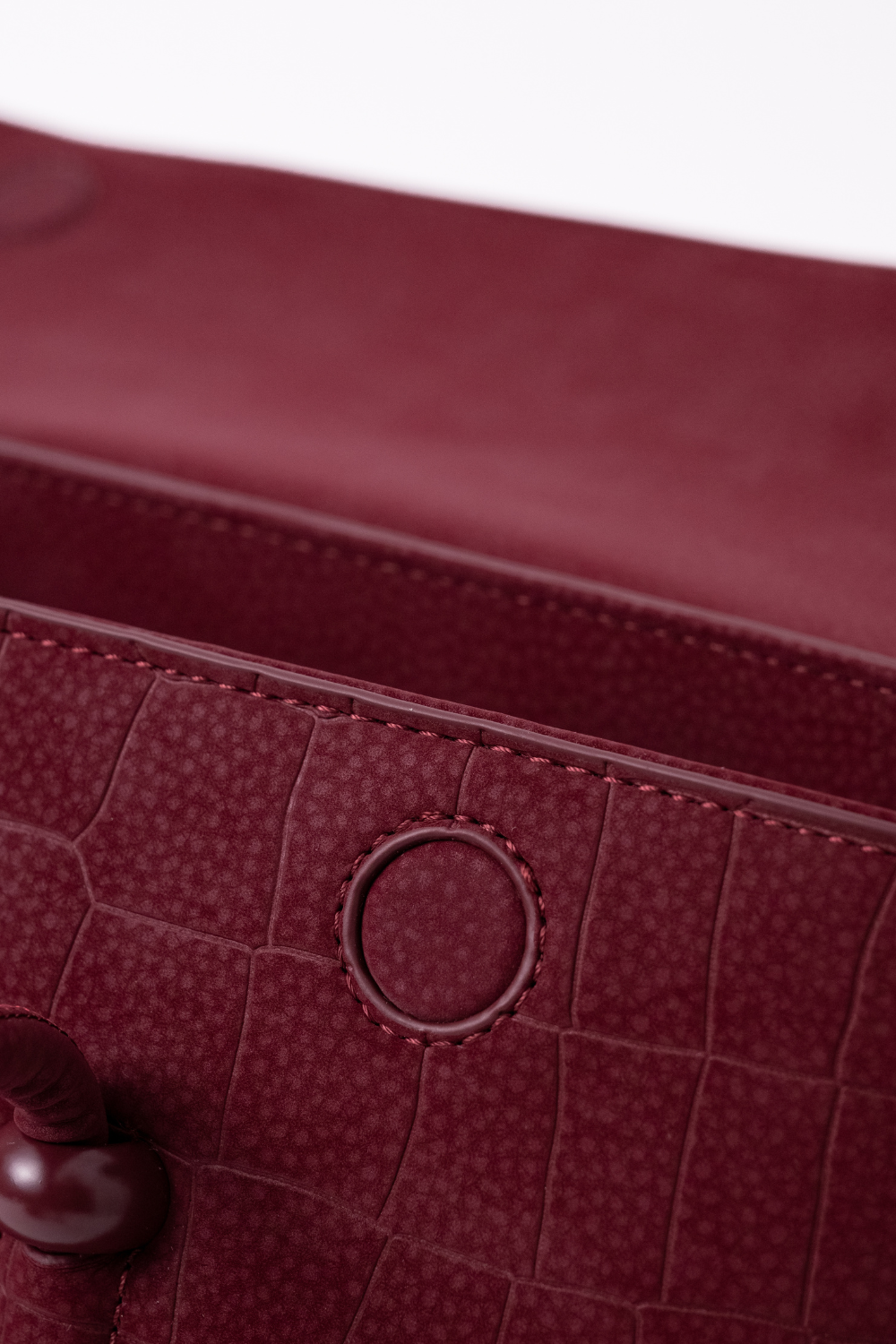 Close-up of a burgundy textured leather bag, showing a crocodile-embossed pattern, a circular metal ring detail, and a dark wooden button on a white background.