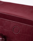 Close-up of a burgundy textured leather bag, showing a crocodile-embossed pattern, a circular metal ring detail, and a dark wooden button on a white background.