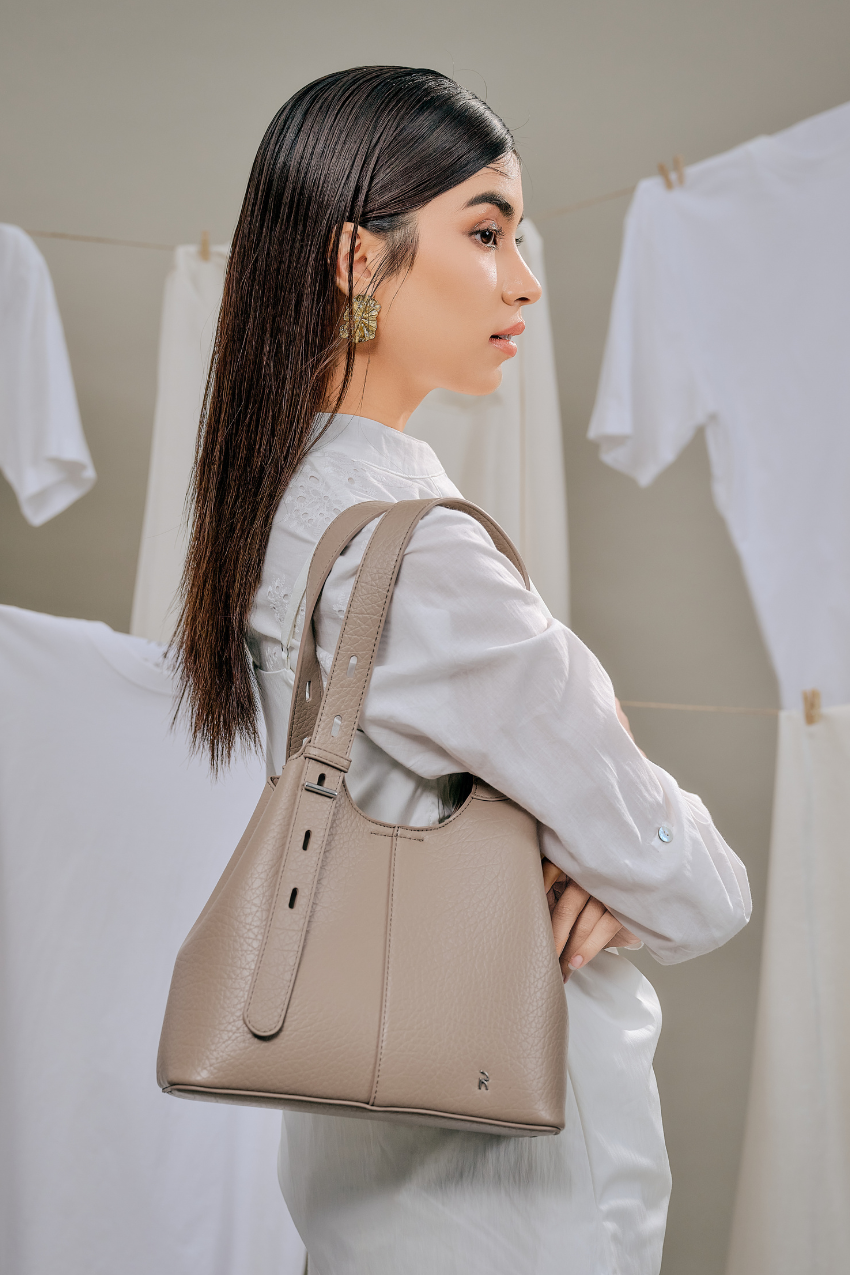 A woman in a white shirt carries a taupe leather shoulder bag with a structured slouchy shape and a small silver logo on the bottom right, set against a background of hanging white shirts.